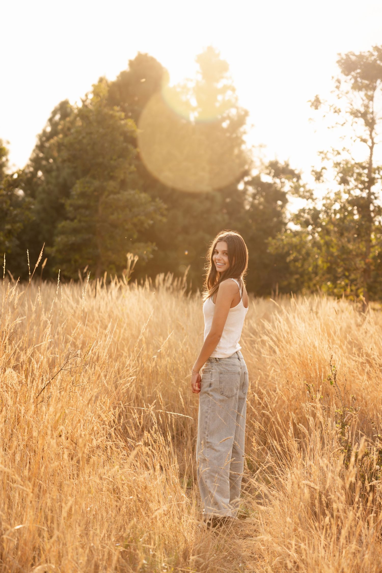 Person standing in golden grass field at sunset with backlit glow and trees in background.