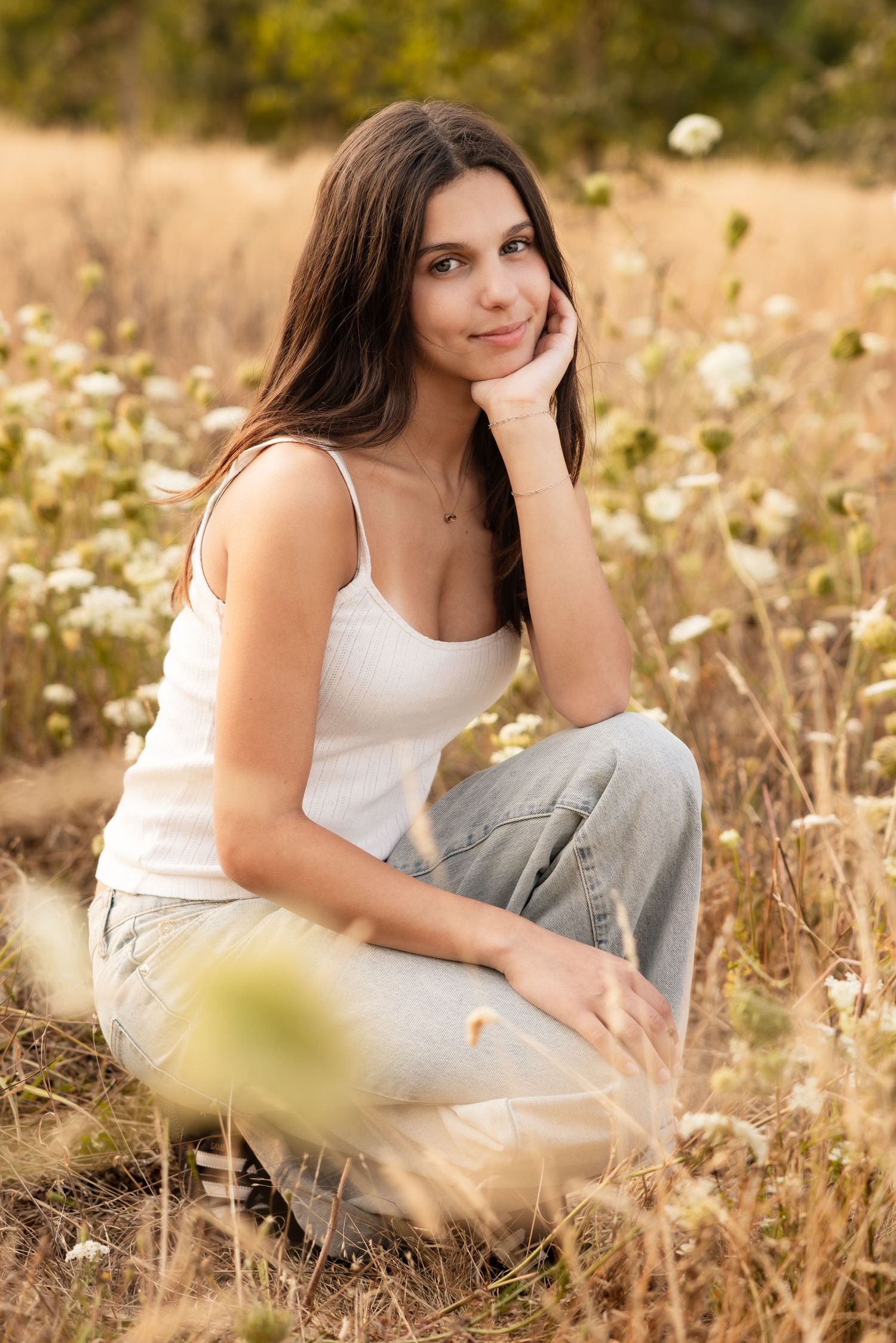 A young woman in a white tank top and jeans poses candidly in a sunlit meadow with wild flowers and golden grass.