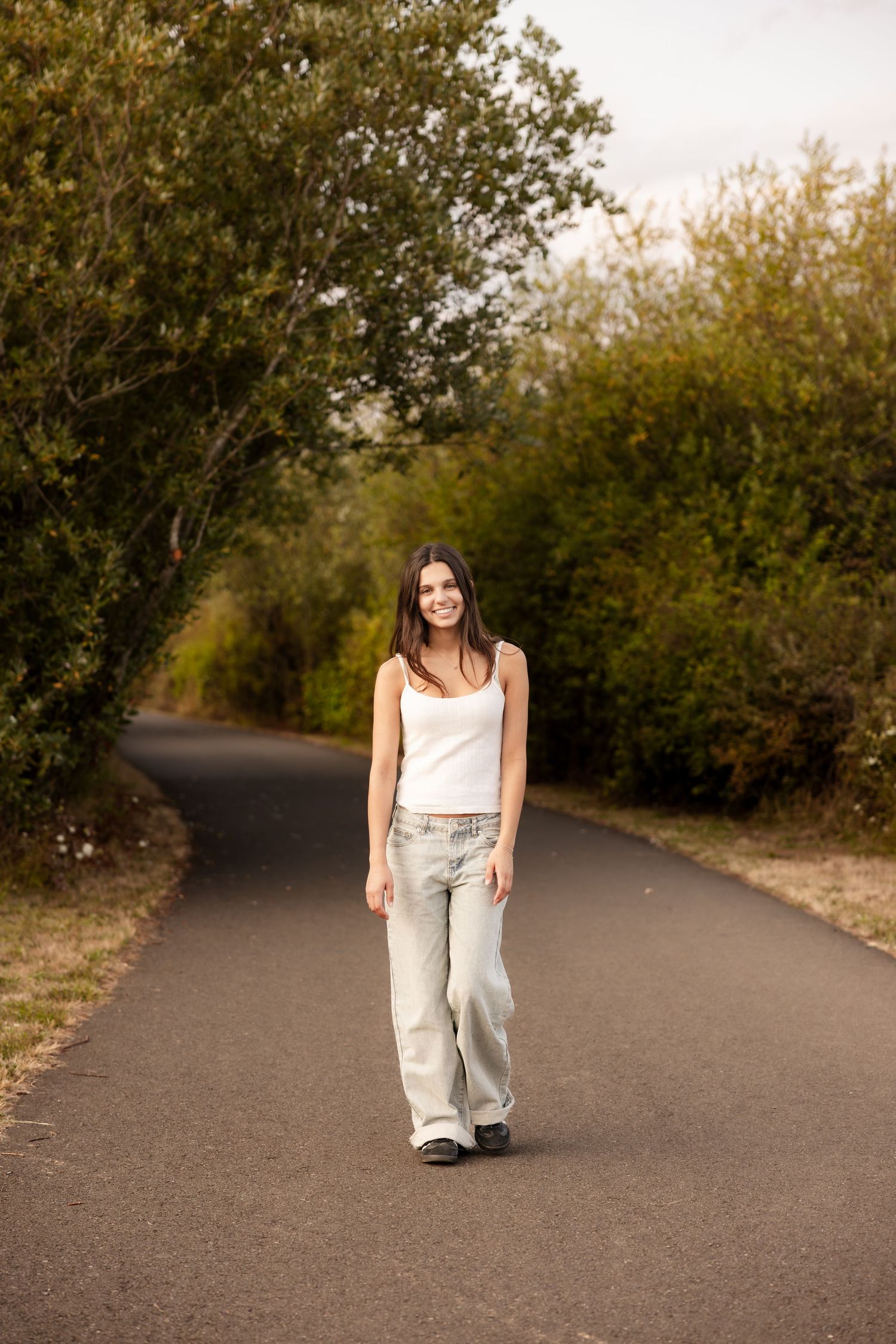 A person in a white tank top and light jeans walks alone on a tree-lined paved path on a sunny day.