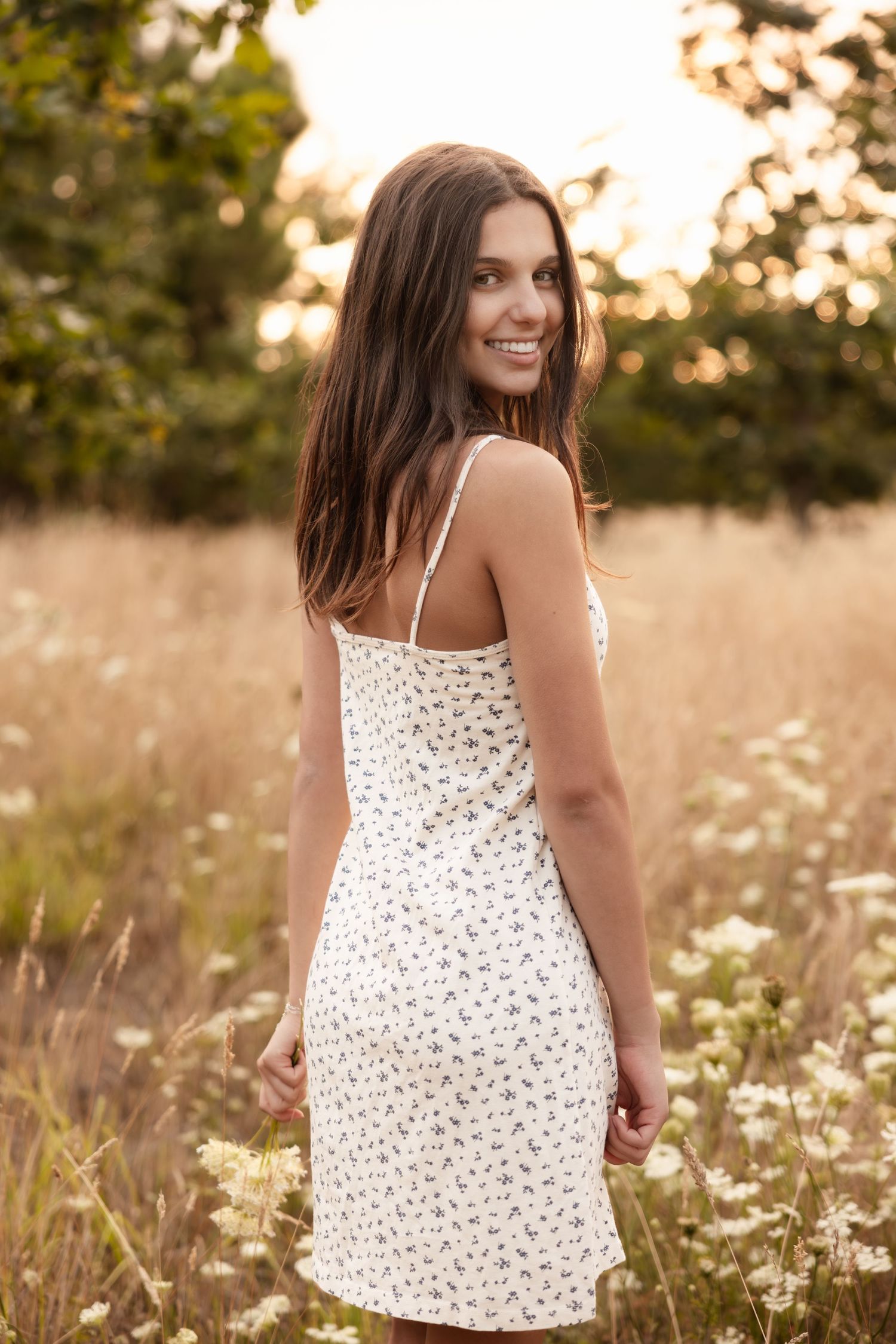 A woman wearing a white floral dress looks over her shoulder while standing in a sunlit field of tall grass.