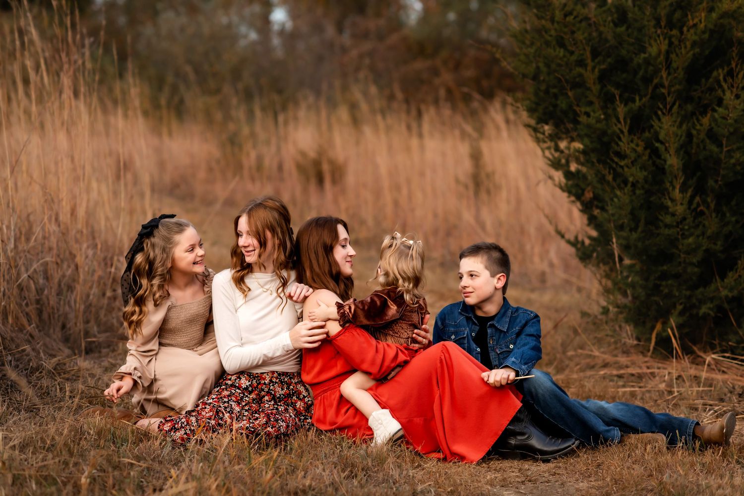 A family group relaxes together on the ground against a backdrop of golden tall grass and evergreen trees.