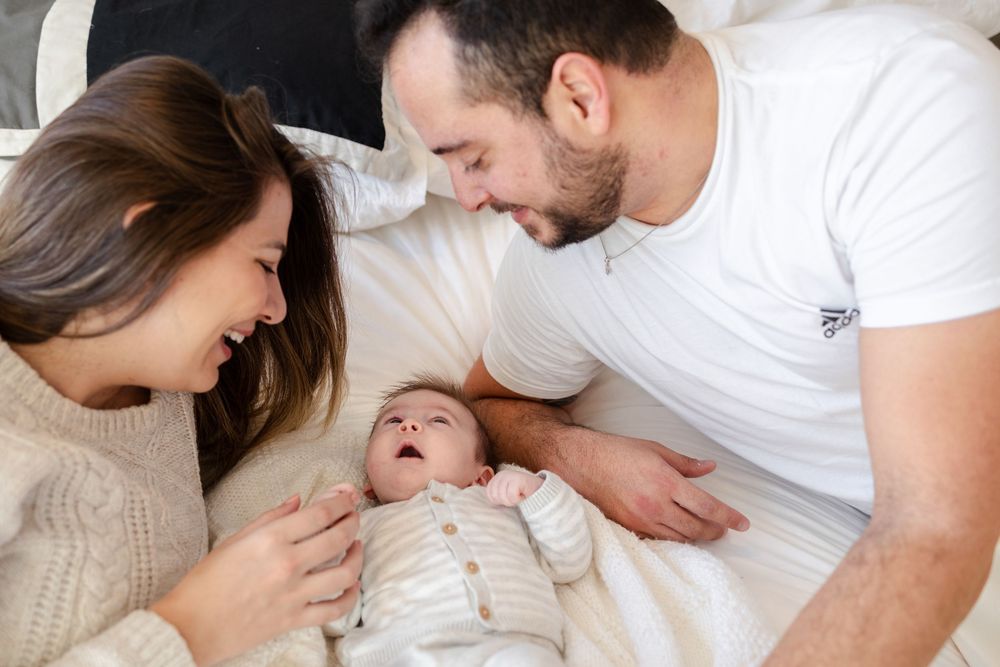 Parents share a joyful moment with their laughing baby while laying together on a white bed.