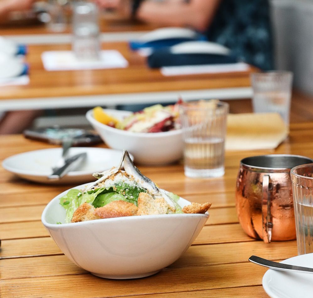 Fresh salad served in white bowl with copper mug on wooden outdoor table.