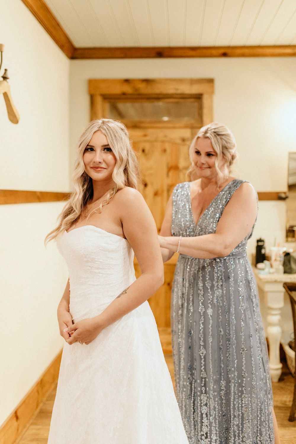 Two people in an elegant room with wooden trim, one wearing a white gown and one in a sparkly silver dress.
