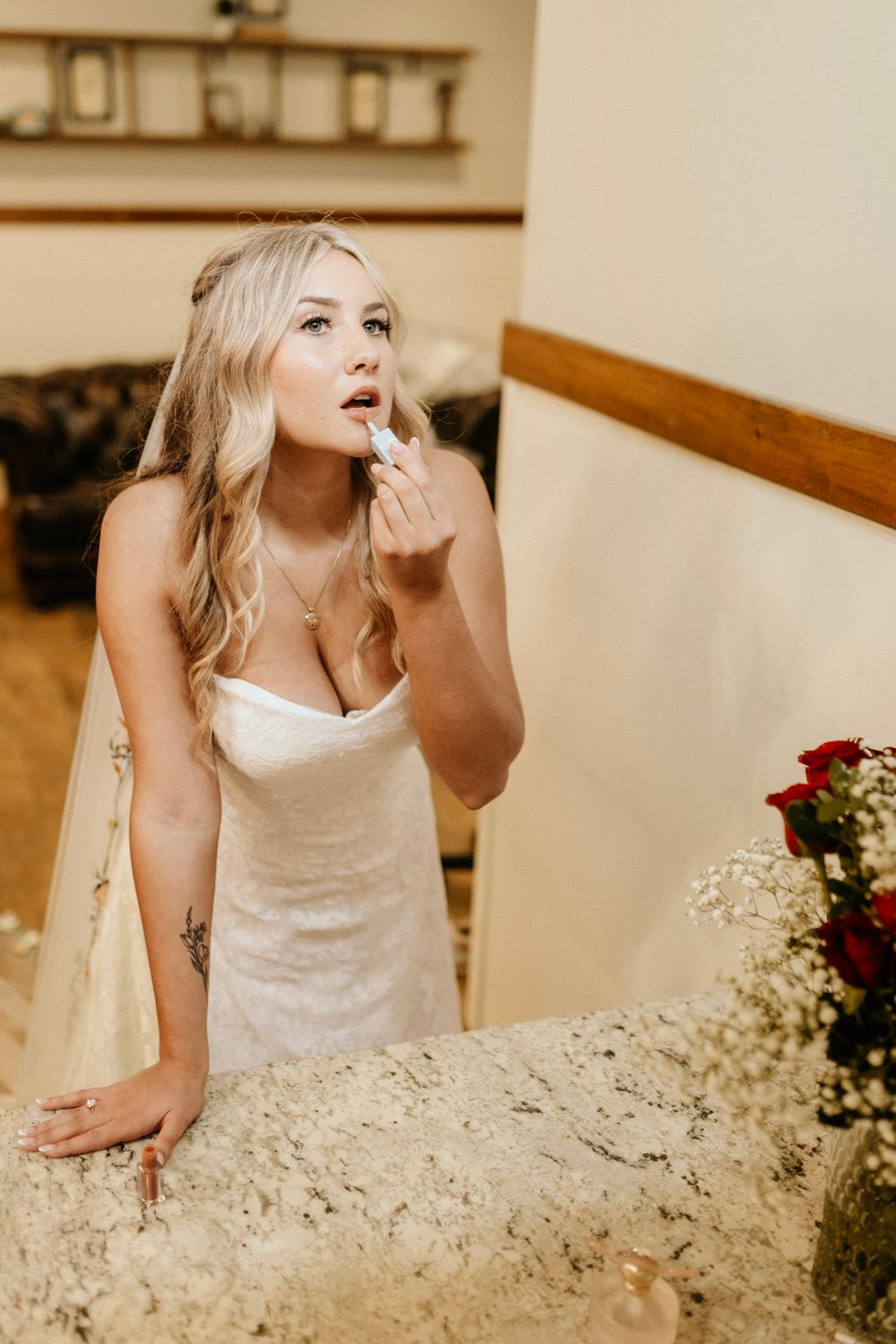 A bride in a white strapless gown applies lipstick while leaning on a rustic surface near floral arrangements.