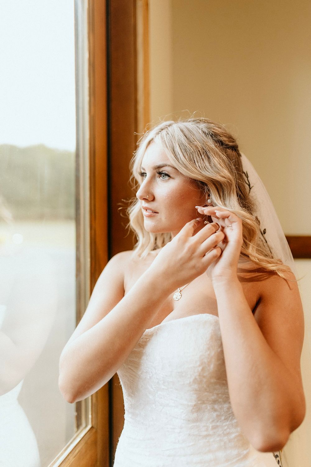 A bride in a white dress adjusts her earrings while standing by a window in natural light.