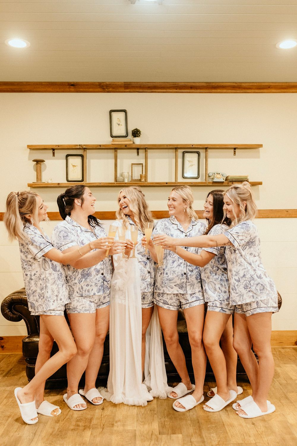 A group wearing matching floral loungewear pajama sets toasting and celebrating around a wedding dress.