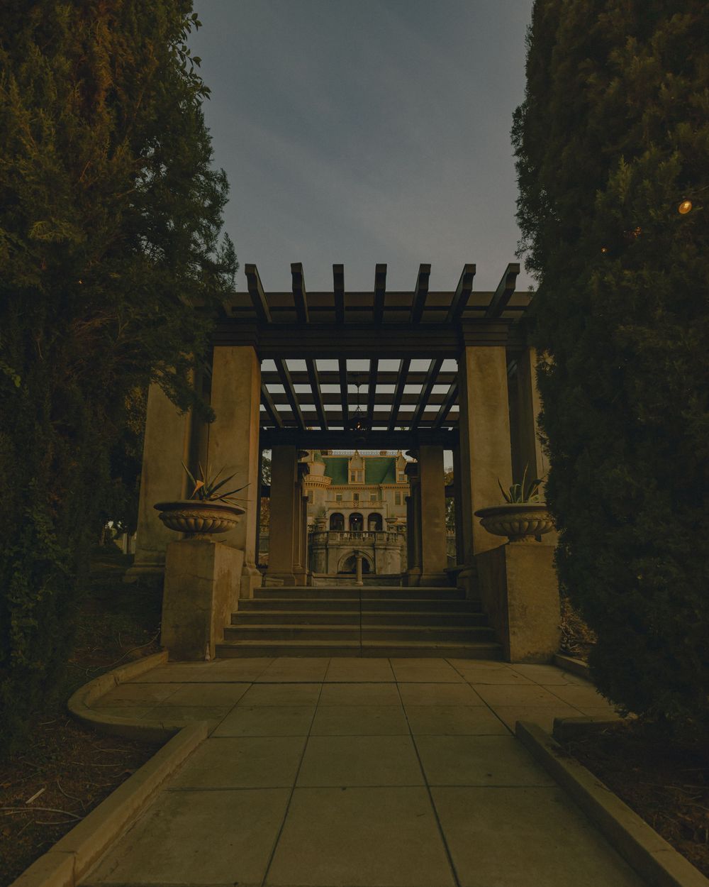 A dark covered walkway with wooden pergola leads to a yellow building entrance between tall trees at dusk