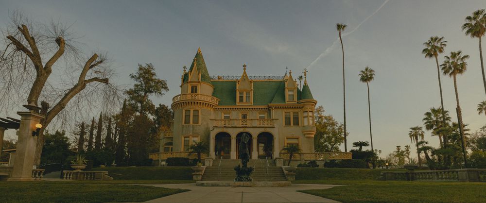 Gothic Victorian mansion Wedding Venue in SoCal with a grand porch and green roof stands majestically among palm trees at dusk