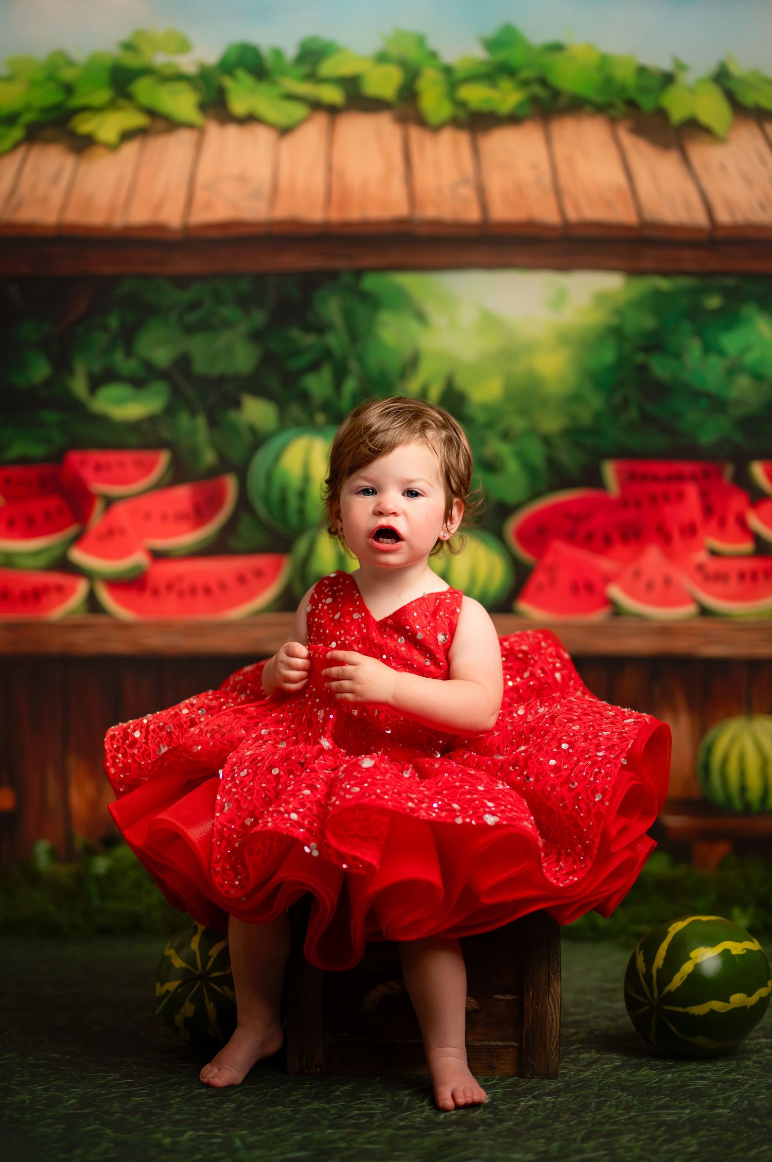 Small child wearing sparkly red holiday dress and headband stands on white pedestal against bright red background.