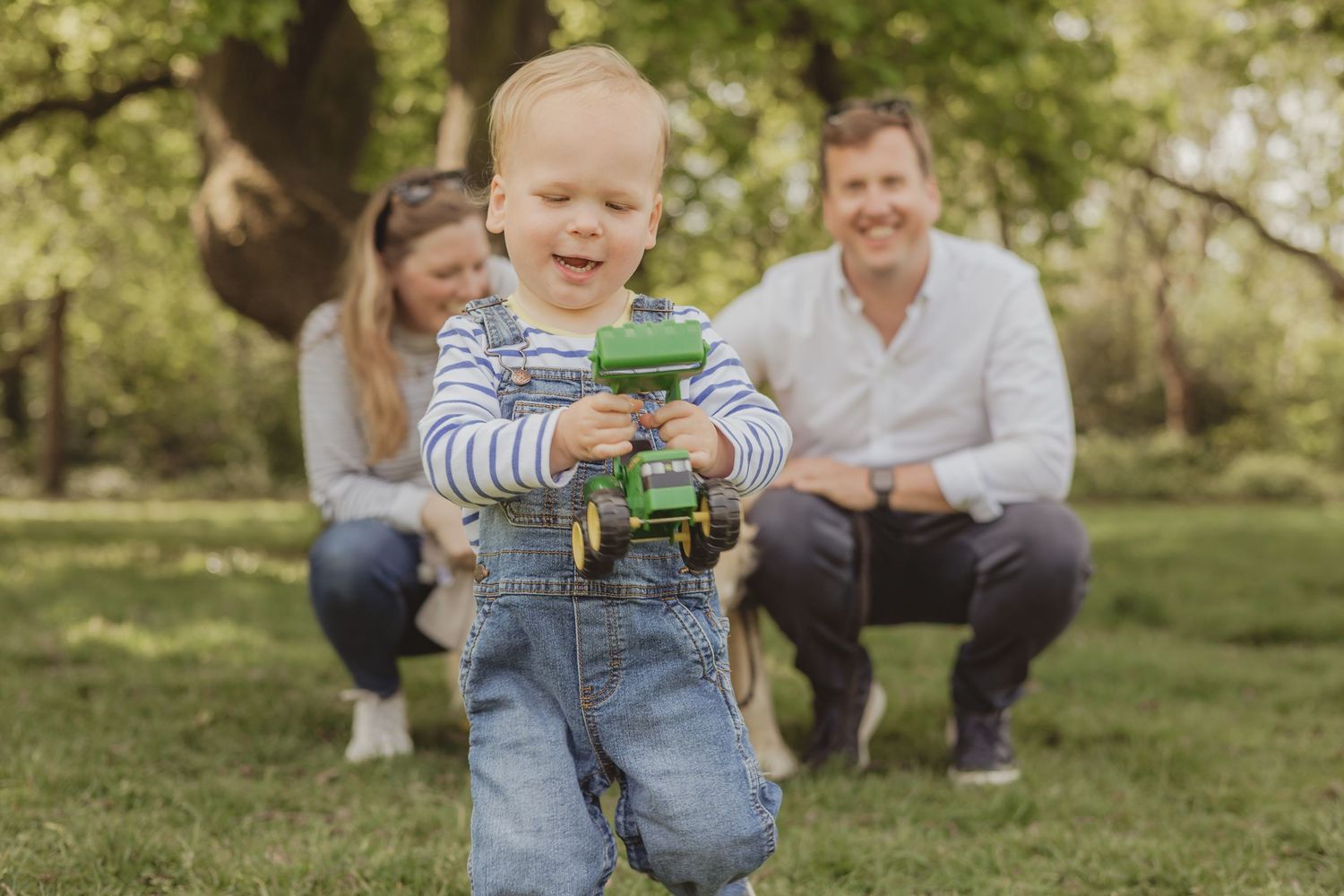 toddler runs towards the camera , holding a green tractor, in a family photoshoot on Wandsworth common