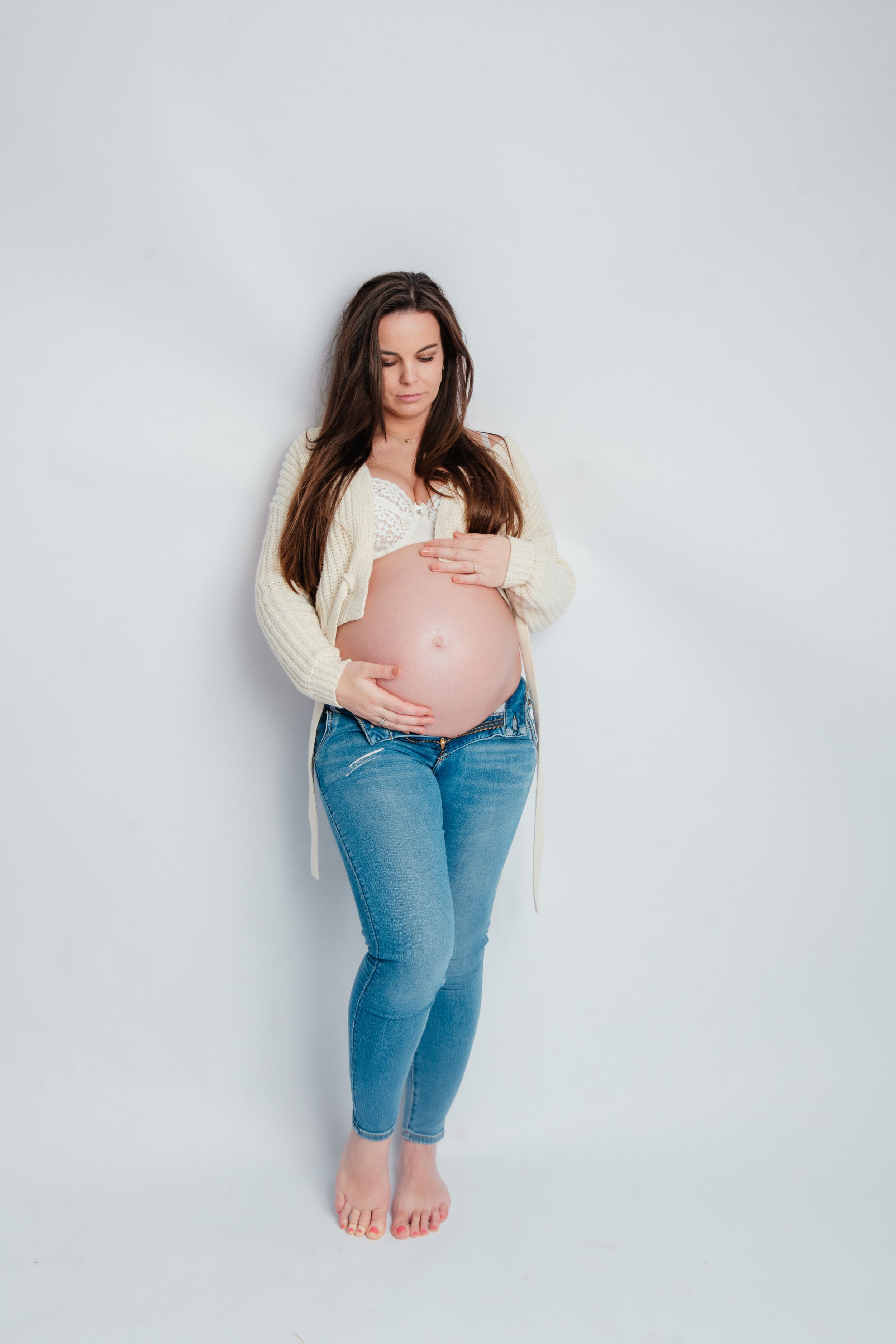 Aanstaande vrouw in jeans en crèmekleurig vest die babybuil wiegt tegen witte muurachtergrond van de fotostudio