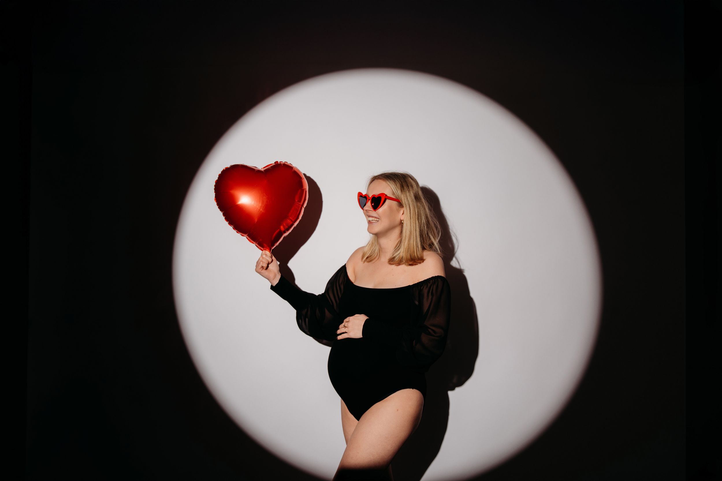 Figuur in een zwarte bodysuit poseert met een rode hartballon tegen een ronde witte spotlight op een donkere achtergrond.