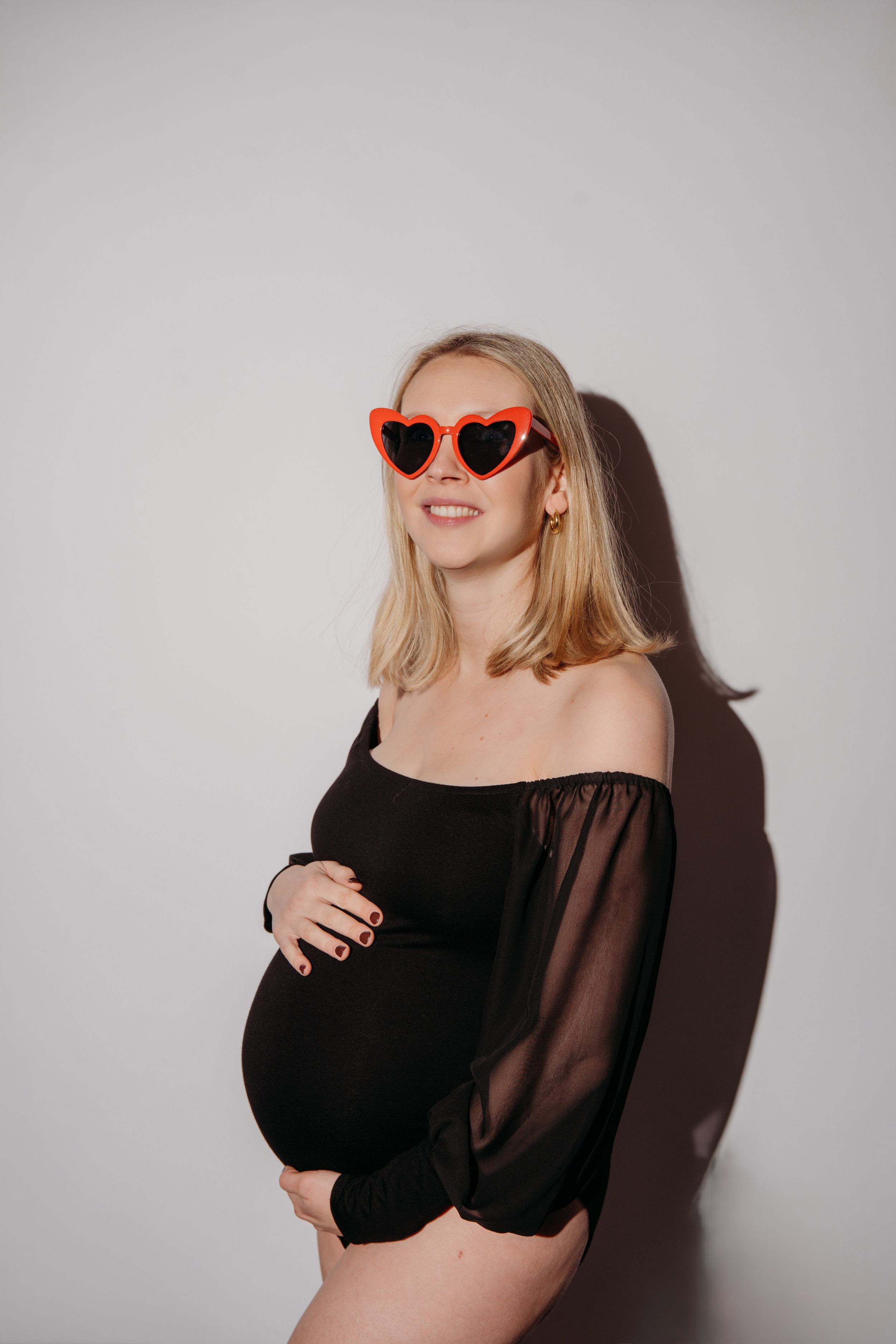 Een zwangere vrouw in een zwarte off-shoulder top en een rode hartzonnebril poseert tegen een witte achtergrond.