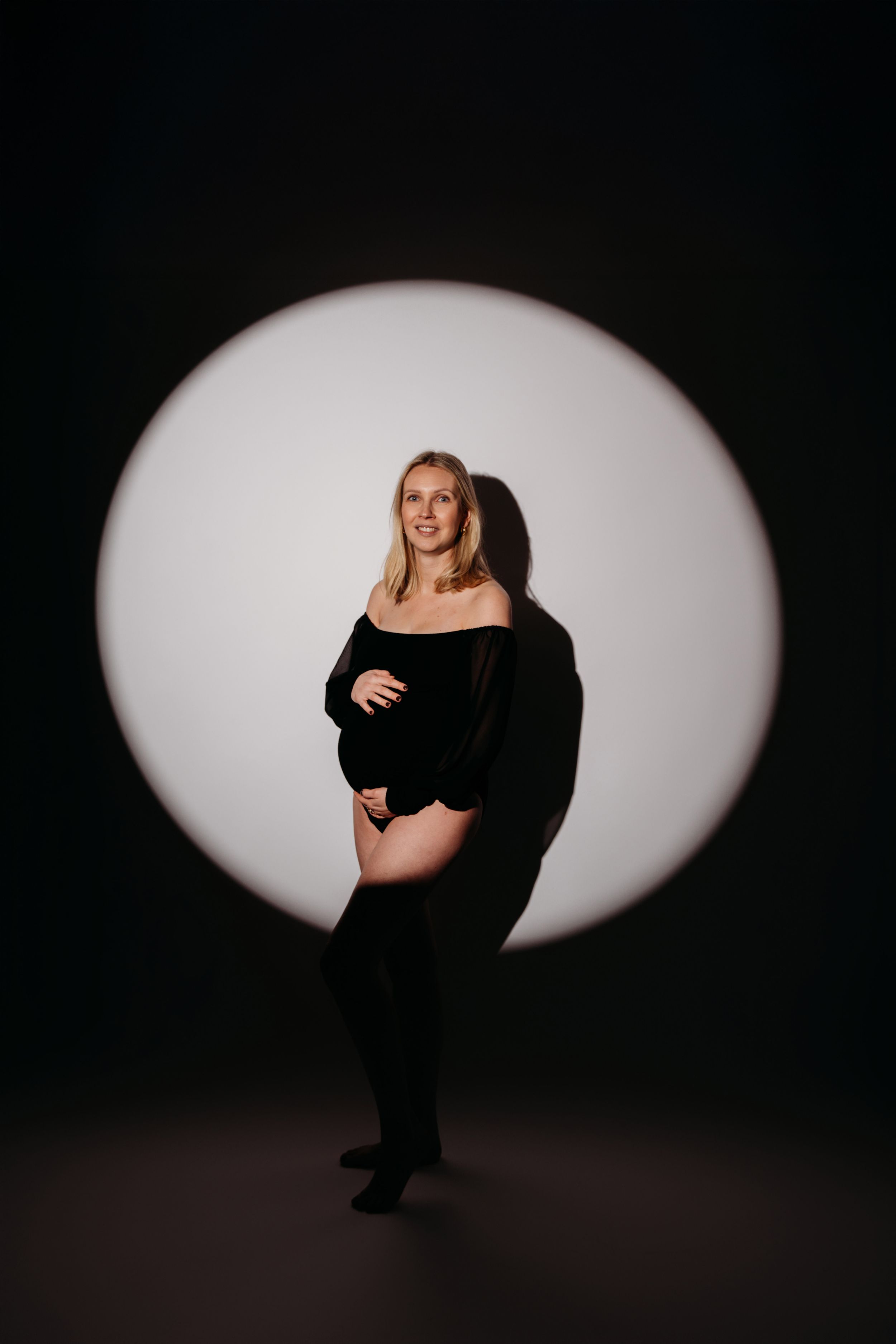Een persoon poseert in zwarte kleding tegen een dramatische ronde witte achtergrond in een donkere studioomgeving.