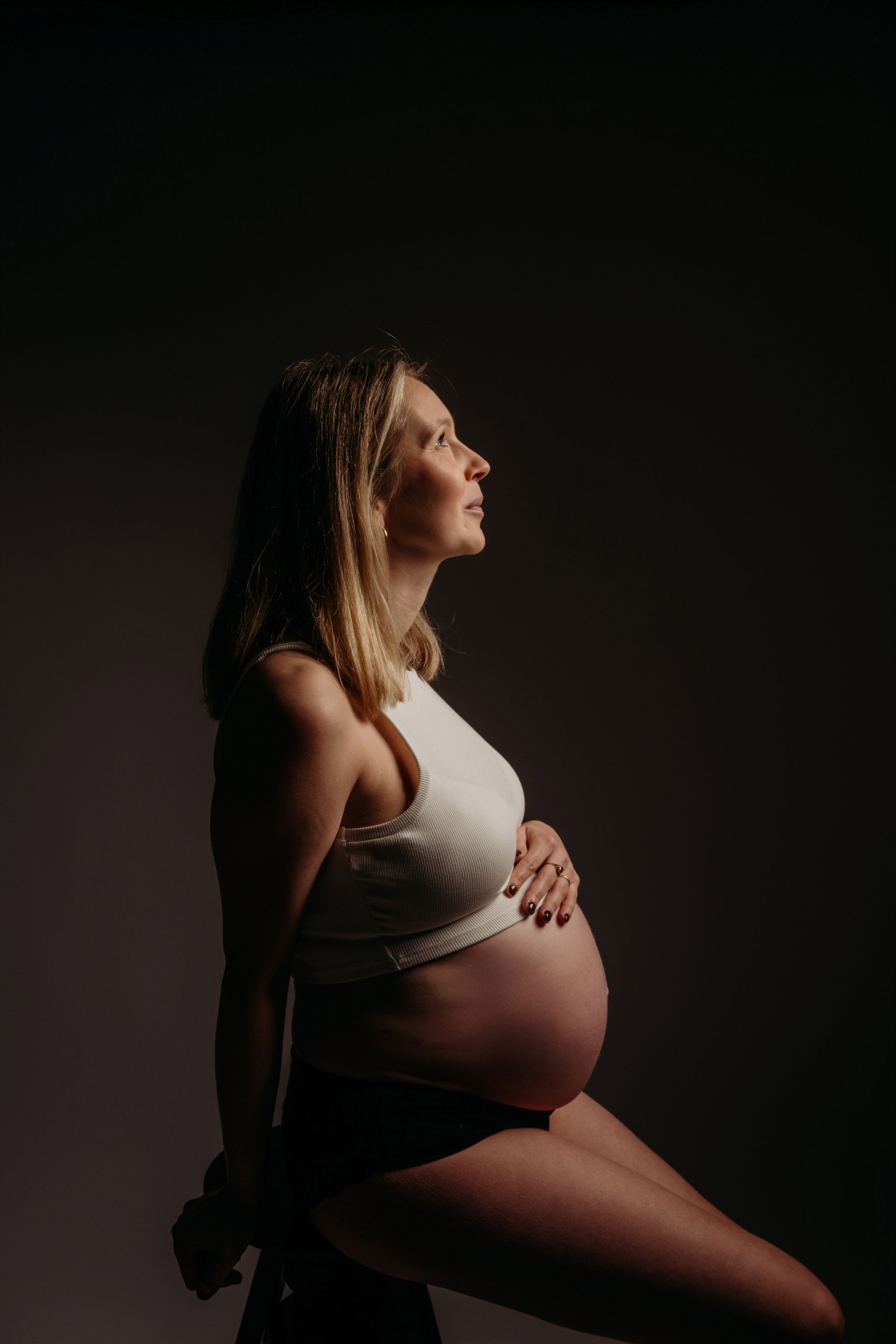 Artistieke zwangerschapsfoto in een donkere studio met aanstaande moeder in witte top die zijwaarts zit in dramatisch licht.