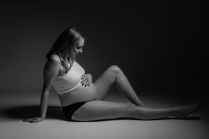 Zwart-wit zwangerschapsfoto van een aanstaande moeder in een crop-top, liggend op de vloer in dramatisch licht.