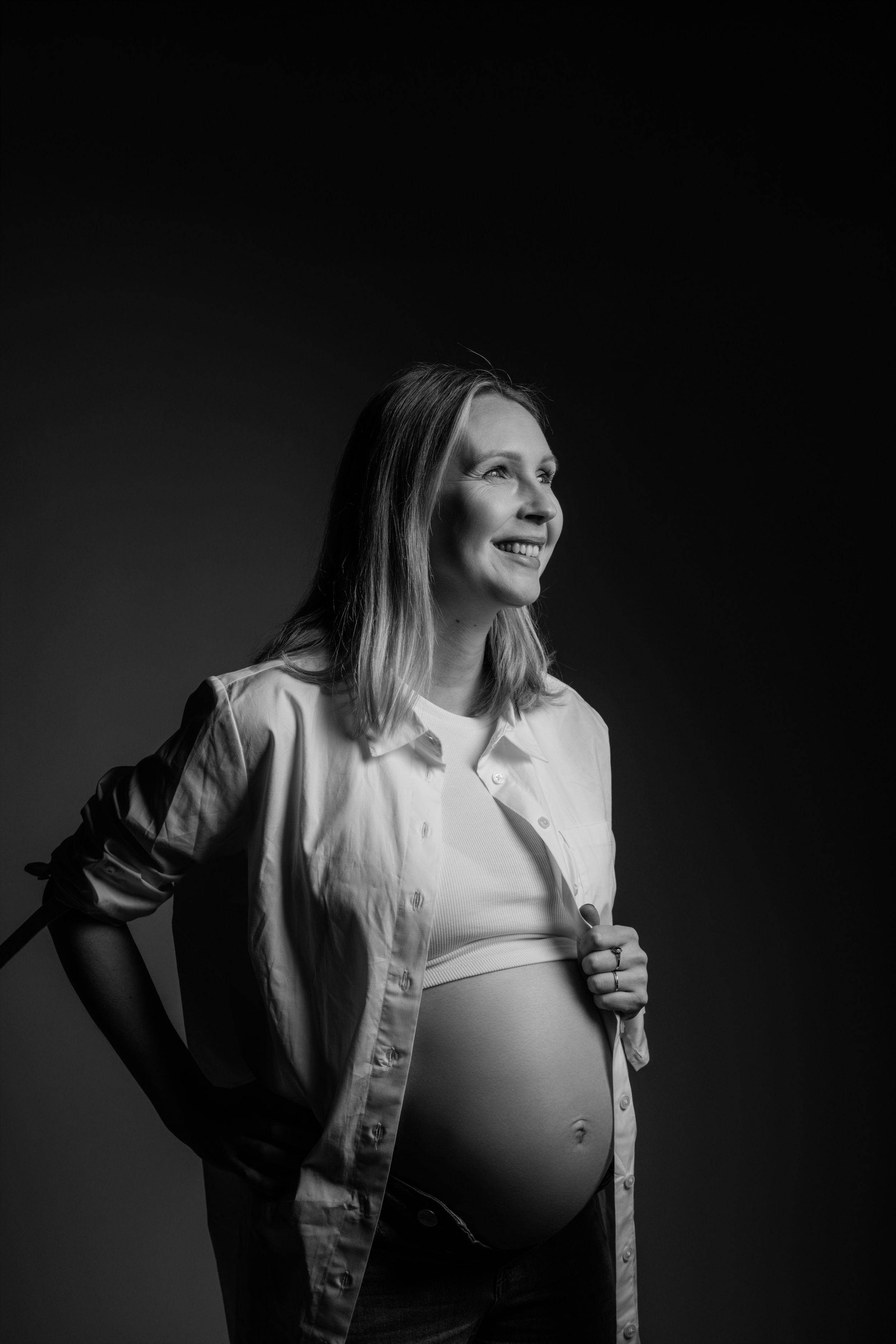 Een zwart-wit portret van een aanstaande moeder in een wit overhemd met knoopsluiting en haar zwangere buik.