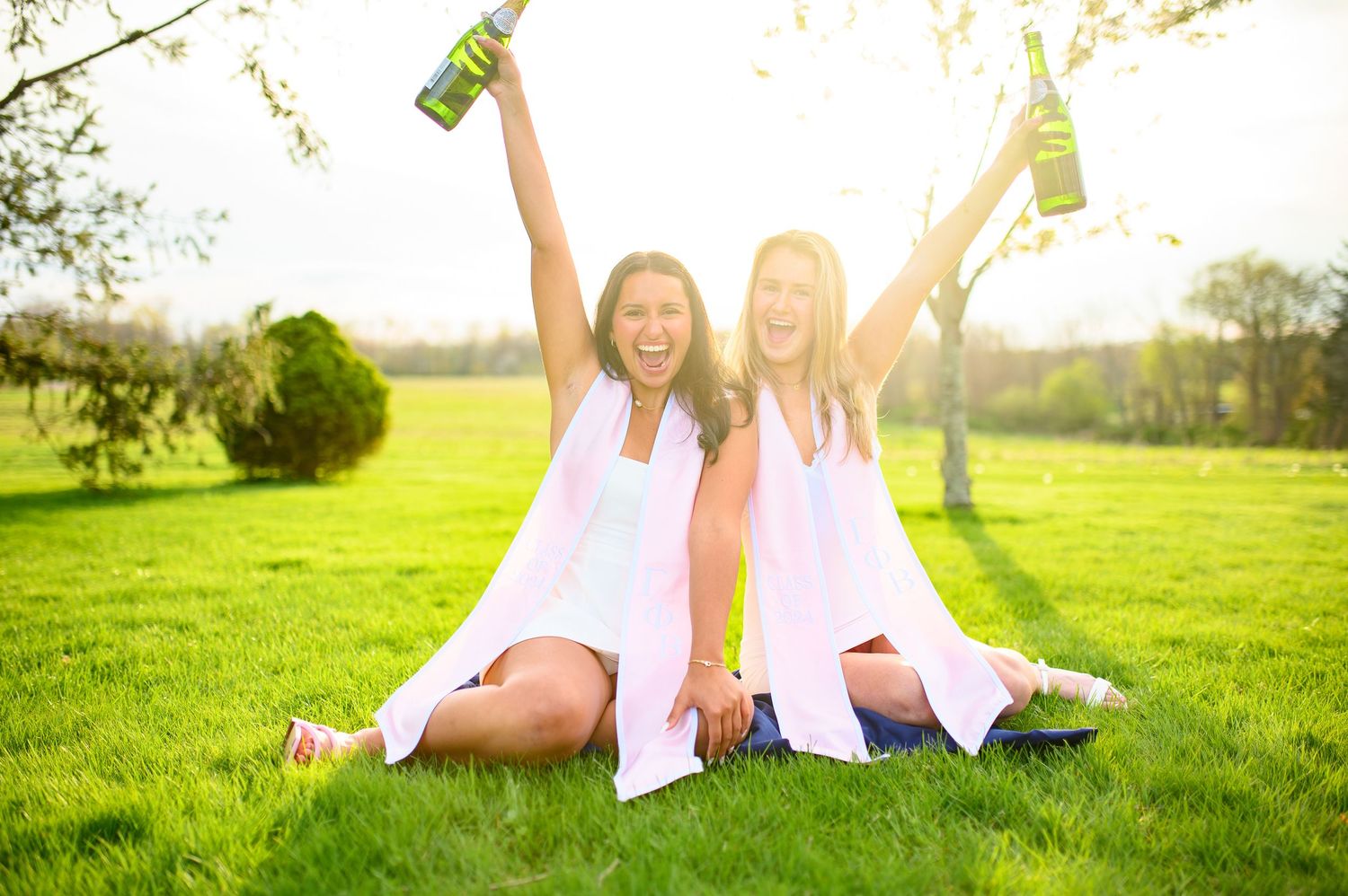 Two people in white dresses sitting on grass holding up bottles while celebrating outdoors in sunlight.