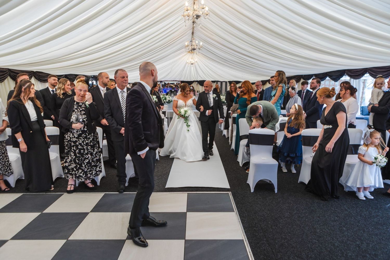 Wedding guests seated in a draped marquee with black and white checkered flooring.