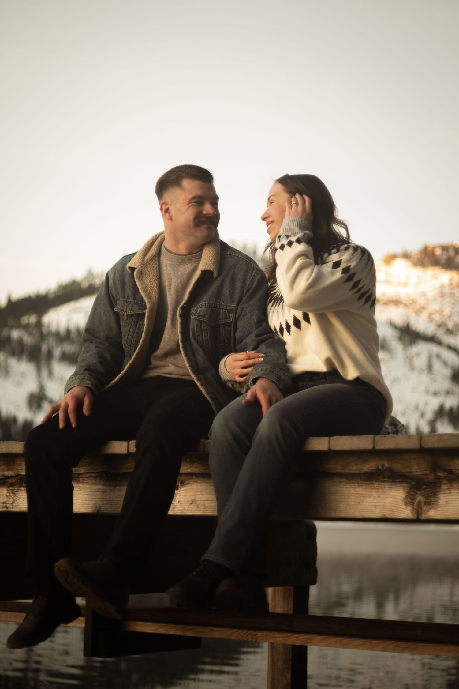 Smiling couple sitting on wooden dock with snowy mountain landscape in the background.