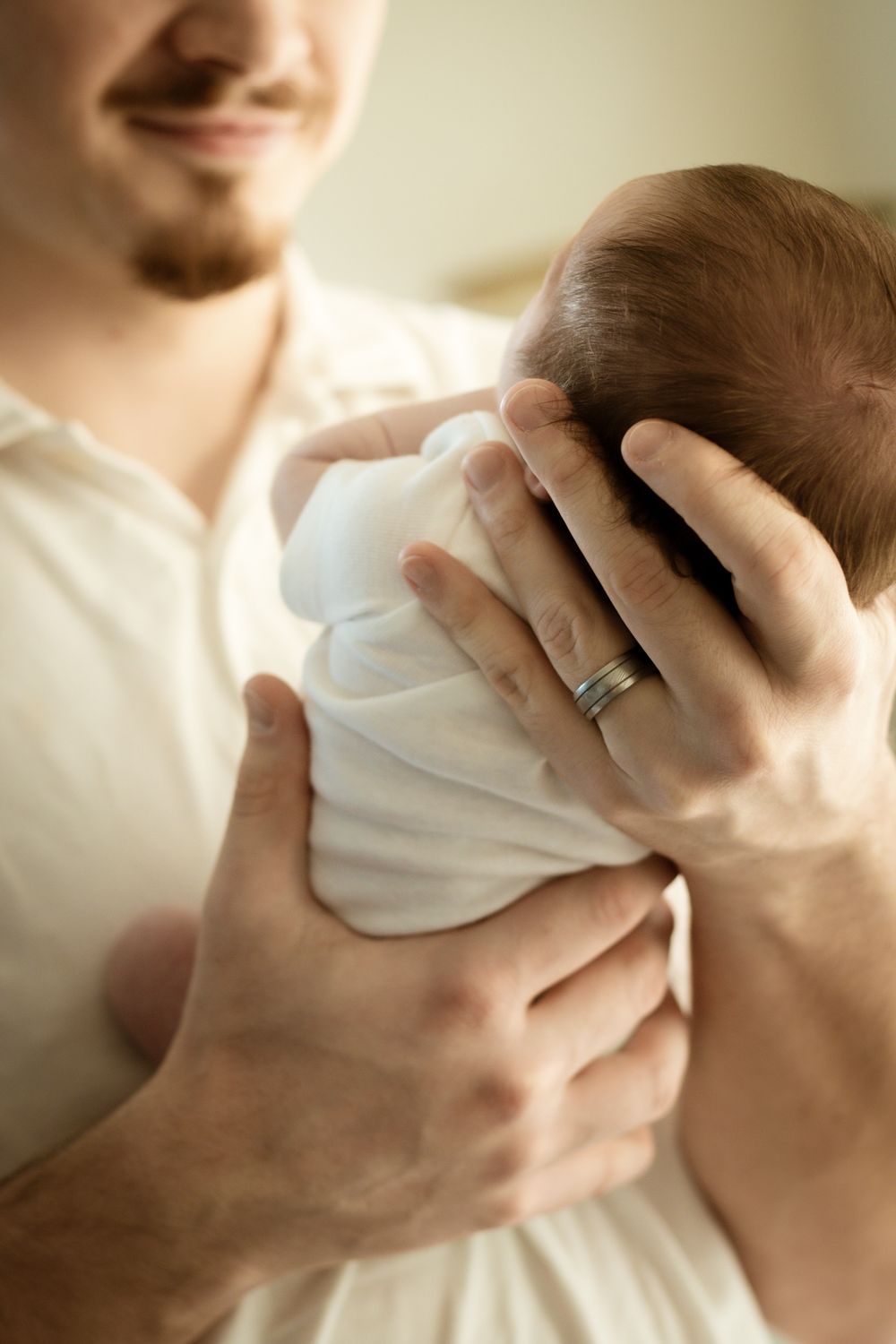Father wearing wedding ring gently cradling newborn baby wrapped in white swaddle in soft indoor light.