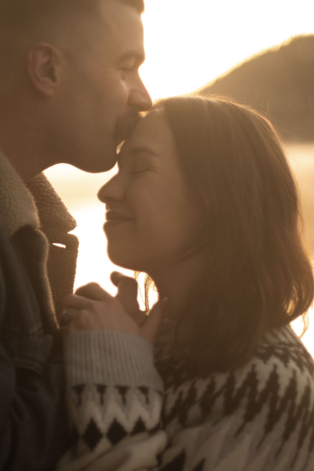 Romantic couple sharing a tender forehead kiss at golden sunset in a warm backlit outdoor portrait.