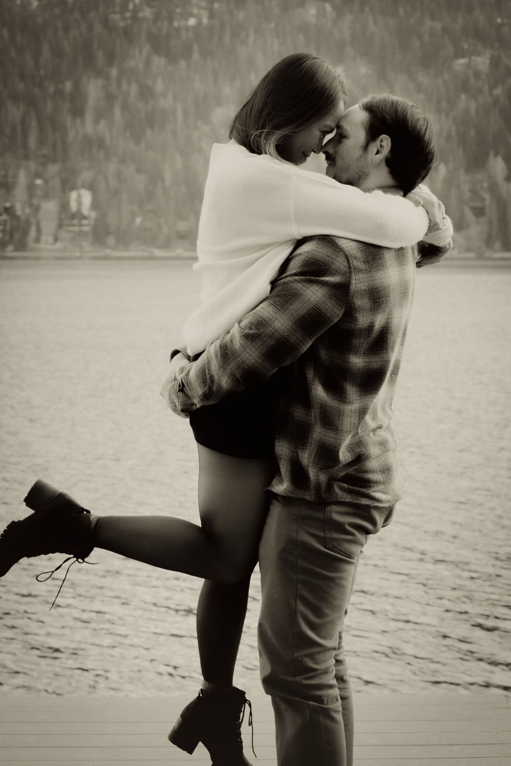 Black and white photo of a couple sharing a romantic kiss by a lake, man lifting woman off the ground.