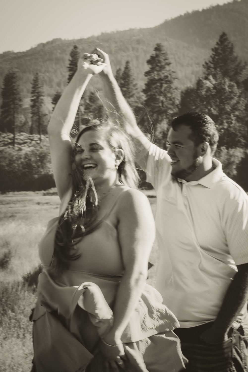 Black and white photo of a joyful couple dancing outdoors with mountain and pine tree scenery behind them.