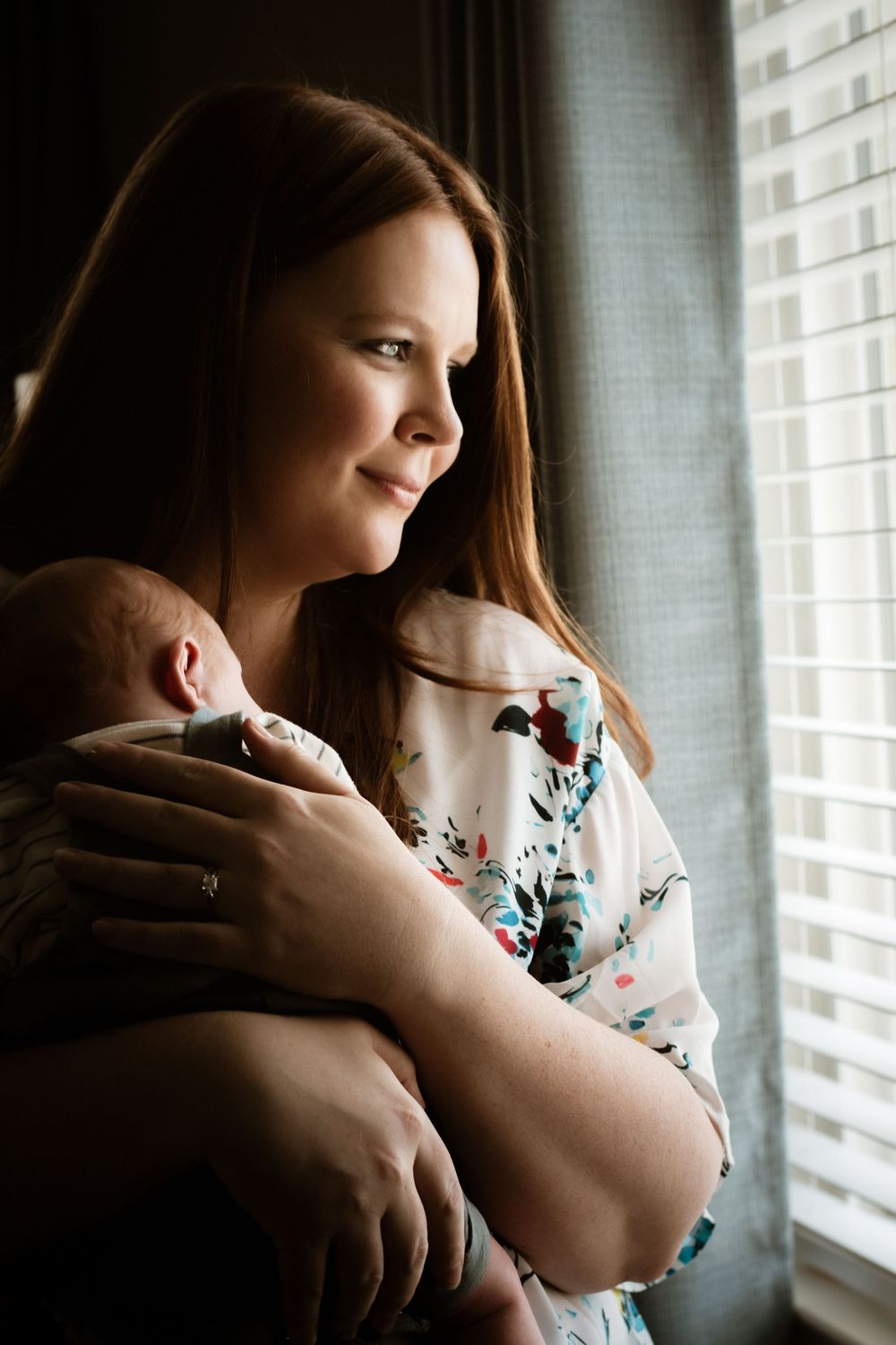 A red-haired mother gazes lovingly at her newborn baby while standing by a window with soft natural light.