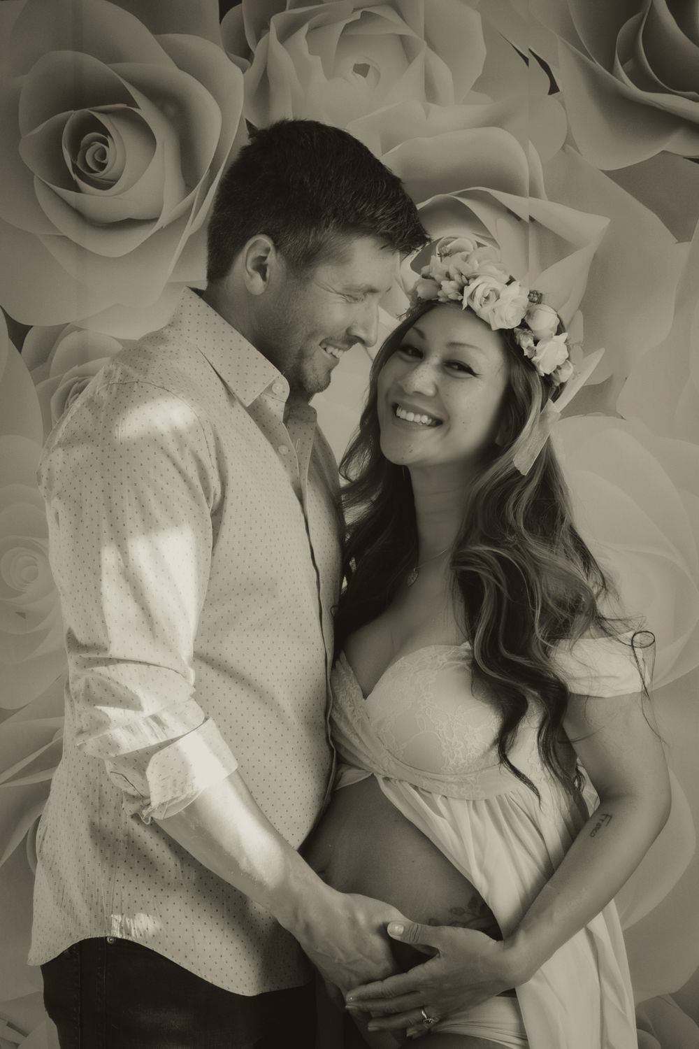 Couple cradles baby bump together smiling in sepia maternity portrait with paper rose backdrop.