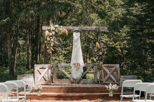 Rustic wooden wedding arch with a white dress hanging in the center and floral decorations in an outdoor forest setting.