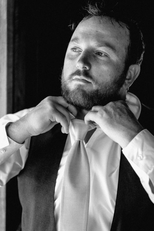 A black and white portrait of a bearded man adjusting the collar of his white dress shirt.