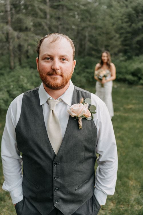 Wedding photo at outdoor venue showing groom in gray vest and white tie with pink boutonniere, bride visible in background.