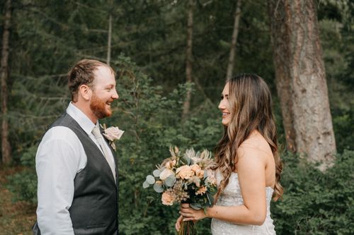 A couple shares a joyful moment in a forest setting during their wedding, with soft floral bouquet and rustic attire.