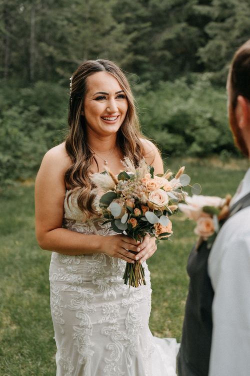 Beautiful wedding photo of the bride smiling outdoors while holding a blush and peach floral bouquet with eucalyptus.