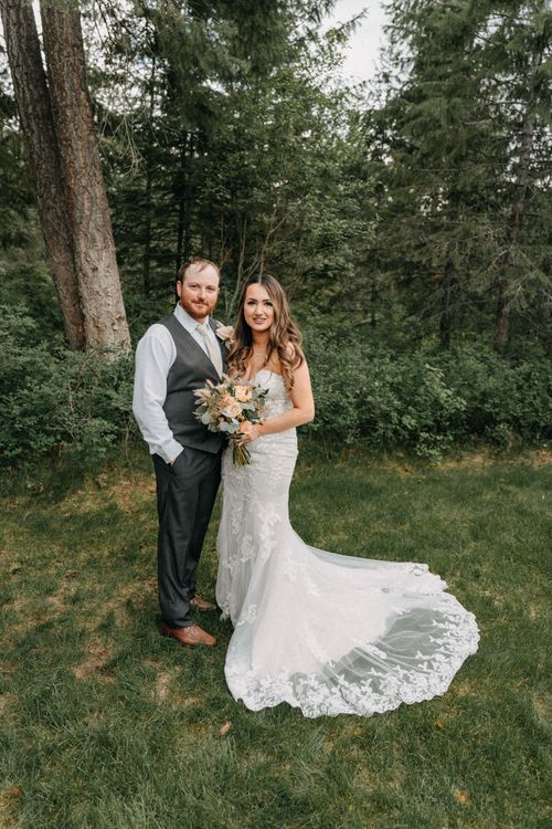 A newlywed couple poses together in a forest setting, with the bride wearing a stunning lace gown with a long train.