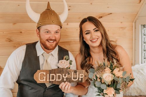 Two people at a rustic wedding holding a 'Giddy Up' wooden sign, captured by wedding videographer Jovan Films.