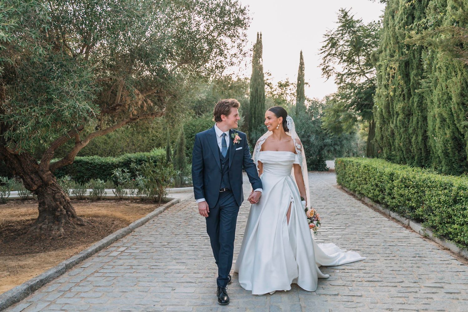 A couple walks along a scenic garden path lined with cypress trees during their wedding celebration at sunset.