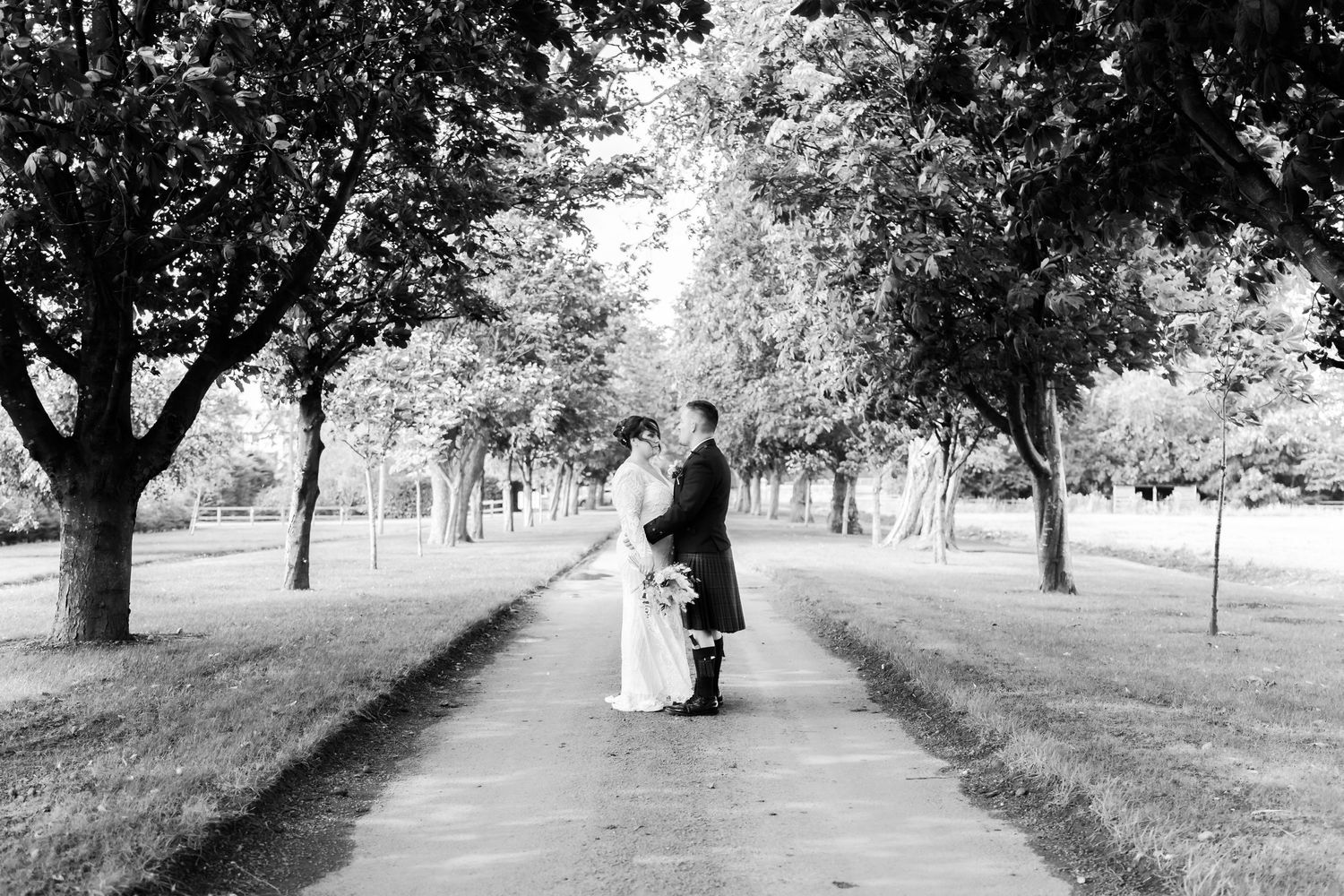A bride and groom stand together on a tree-lined pathway in black and white photography.