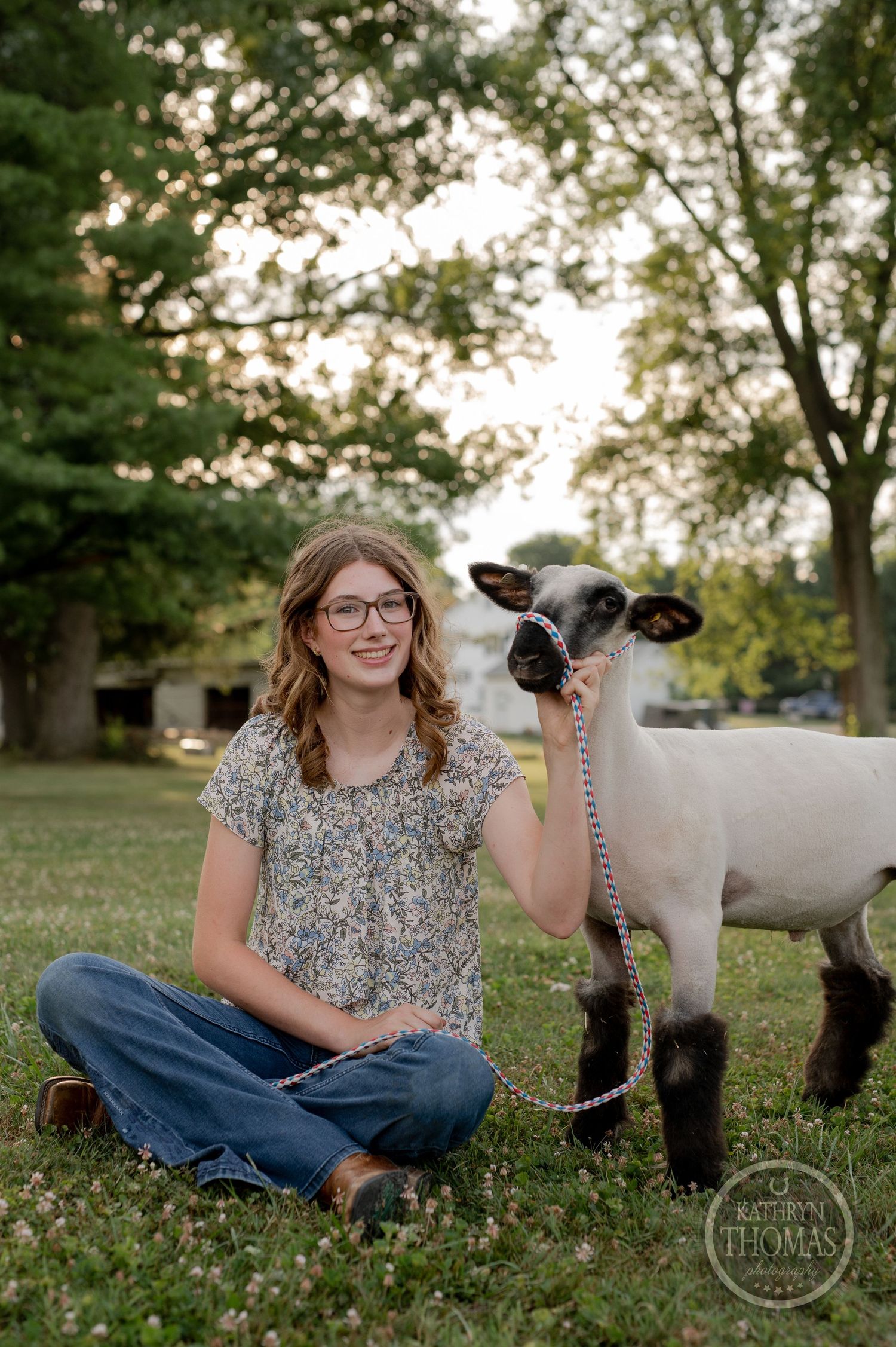 A person in a floral top sits cross-legged on grass next to a show lamb in a pastoral setting.