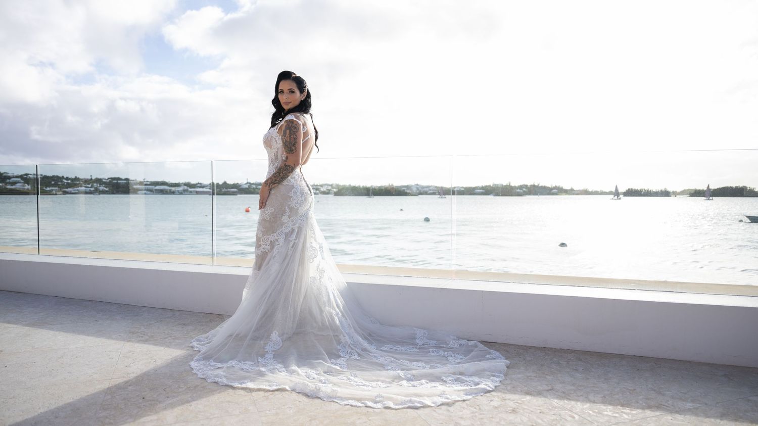 A bride in a flowing white wedding gown poses on a balcony overlooking the ocean.