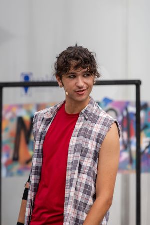 Person wearing a red tank top and sleeveless plaid shirt smiles while posing against colorful background.