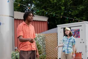 Two people stand in a backyard garden setting with lattice fencing and string lights visible in the background.