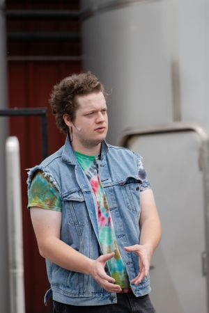 A performer in tie-dye shirt and denim vest gestures during an outdoor scene.