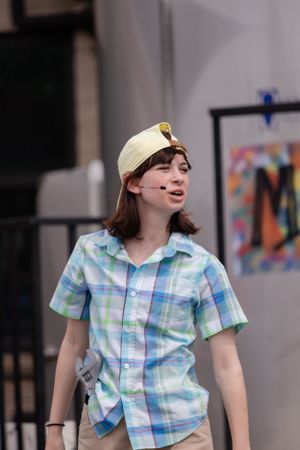 Person in a plaid short-sleeve shirt and beige cap looks to the side while standing outdoors.