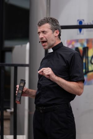 A person in clerical attire speaks animatedly while holding a device outdoors.