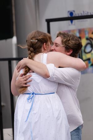 Two people share an emotional embrace, with one wearing a white dress with blue ribbon and the other in casual attire.