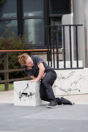 Person in dark clothing slumps dramatically on white marble ledge outdoors.