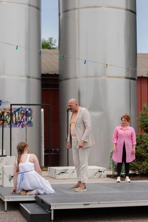 Three performers stand on a gray stage platform in front of large industrial storage tanks with graffiti.